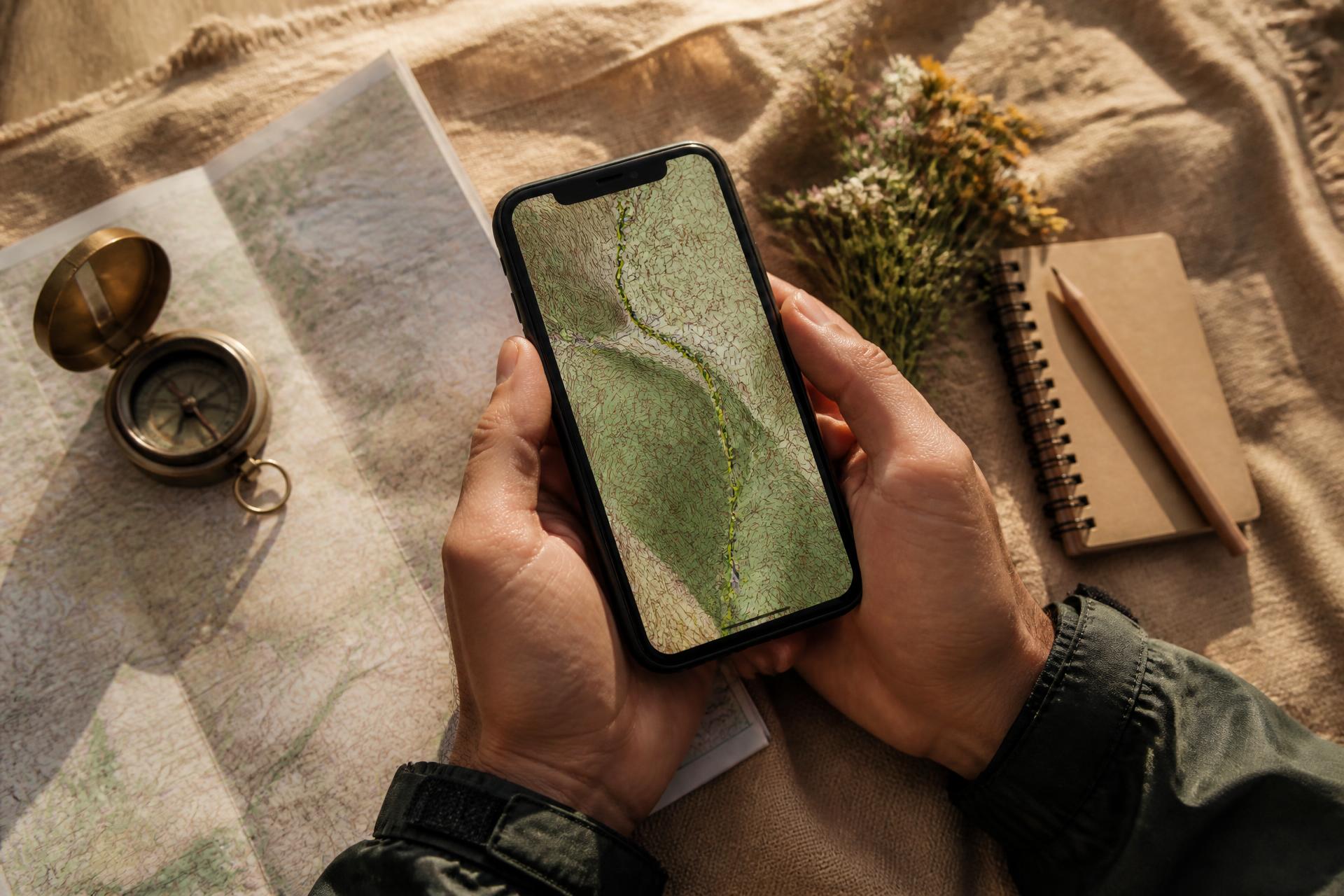 A hiker's hands holding a phone with a topographic offline trail map, beside a brass compass and folded paper map
